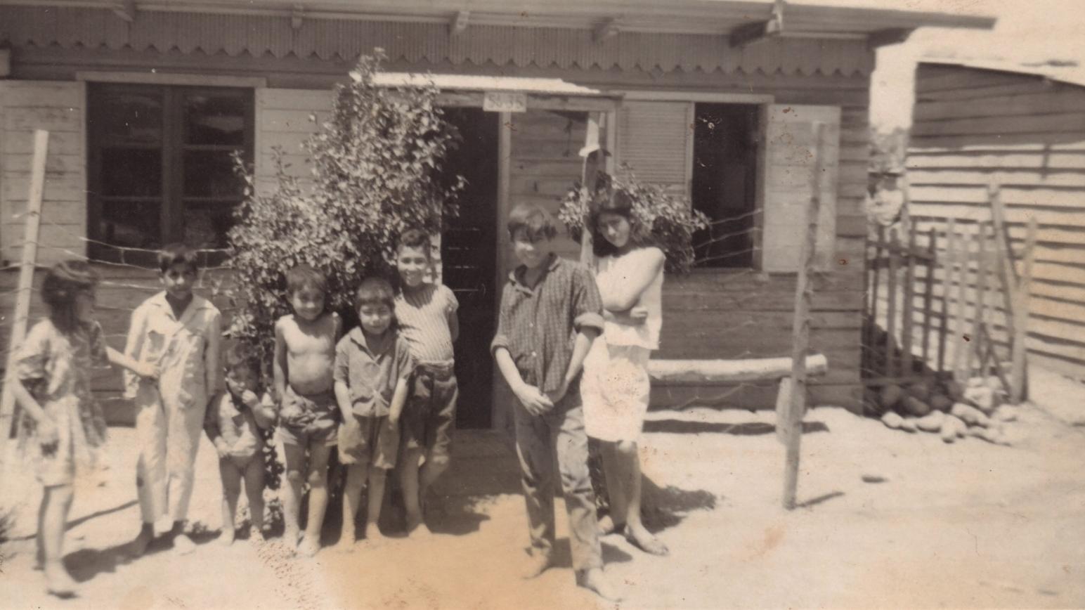 Foto en tonos sepia que muestra a un grupo de niños frente a una casa pequeña de madera