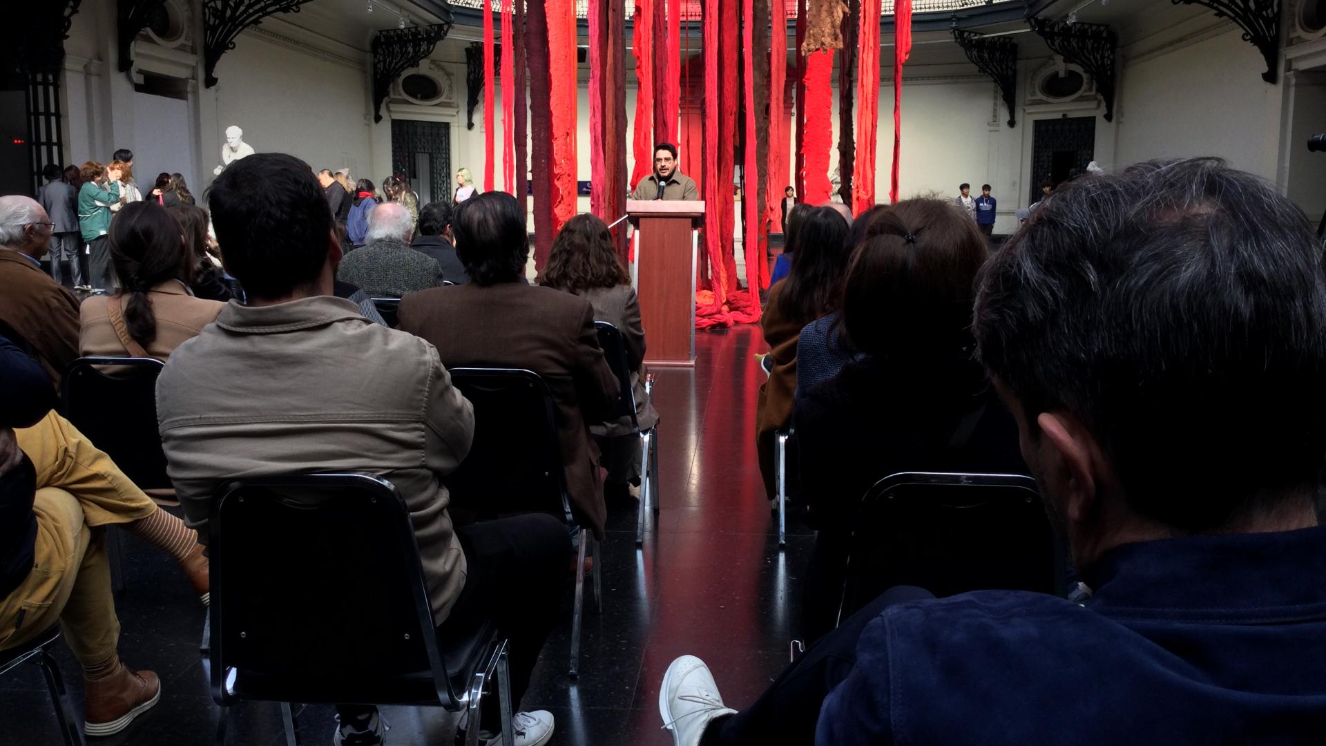 El curador Sebastián Vidal Valenzuela dando un discurso, detrás de él se pueden ver telas colgadas de colores rojo y café. Frente a él hay personas sentadas observandolo.