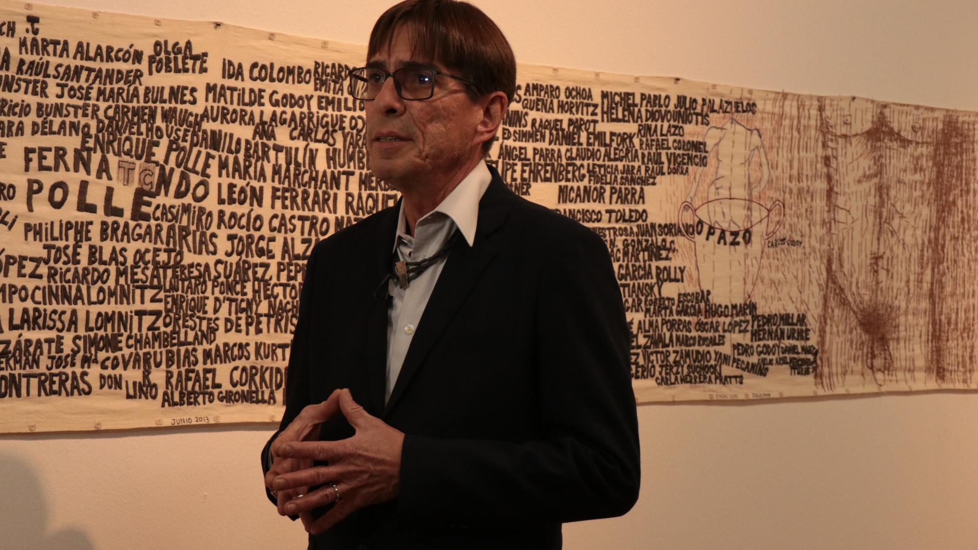 Hombre con lentes, detras de el se encuentra una pieza bordada con hilo negro