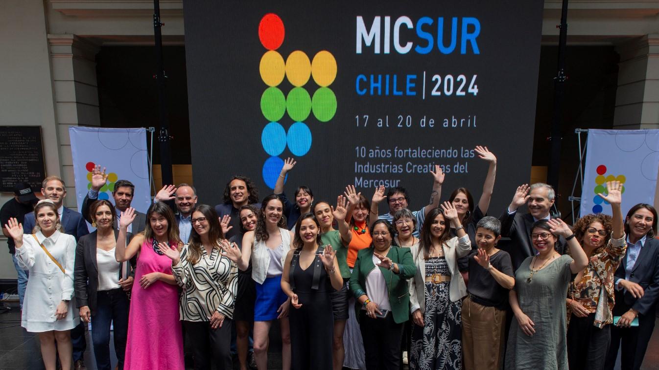 Grupo de personas saludando con las manos en alto, frente a un cartel con el texto "MICSUR Chile 2024"