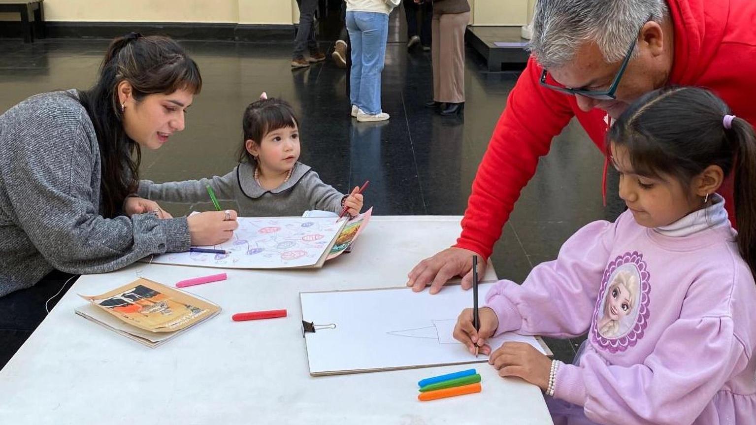 Fotografía de dos niñas dibujando, acompañadas de adultos
