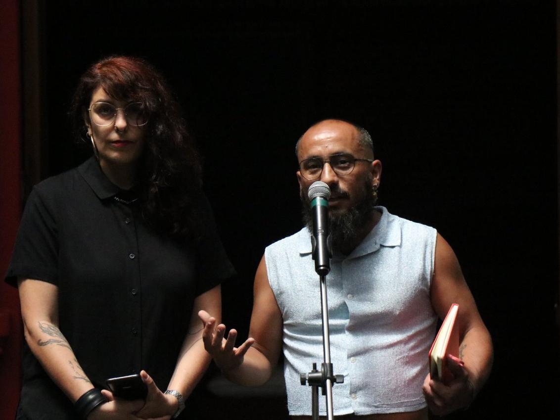 Fotografía de una mujer con blusa negra y un hombre con polera blanca, hablando frente a un micrófono
