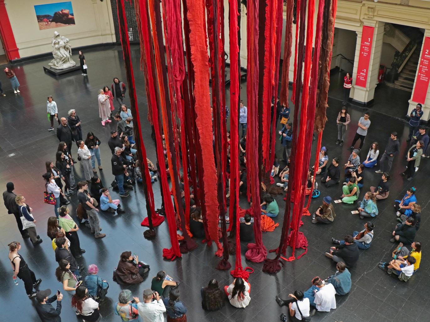 Fotografía de grupo de personas en el hall del Museo, sentadas alrededor de instalación colgante en tonos rojos