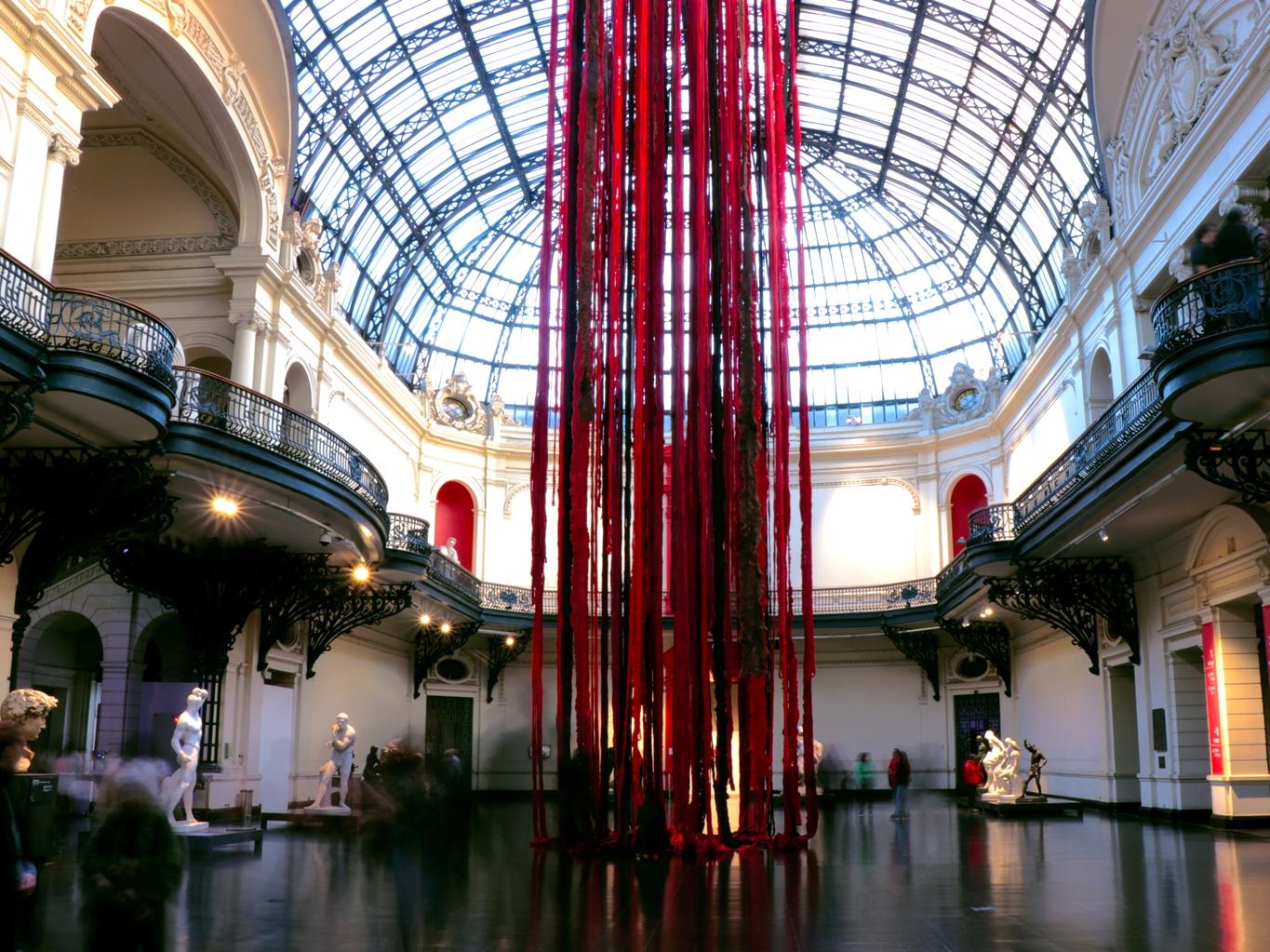 Fotografía del hall del Museo con  obra que consiste en lanas colgantes en tonos rojos