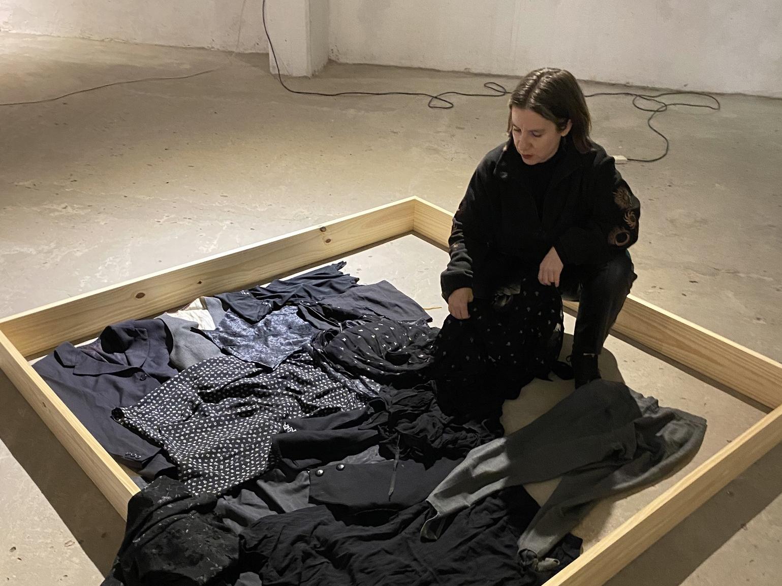 Fotografía de mujer junto a varias prendas de vestir negras, dispuestas en el suelo