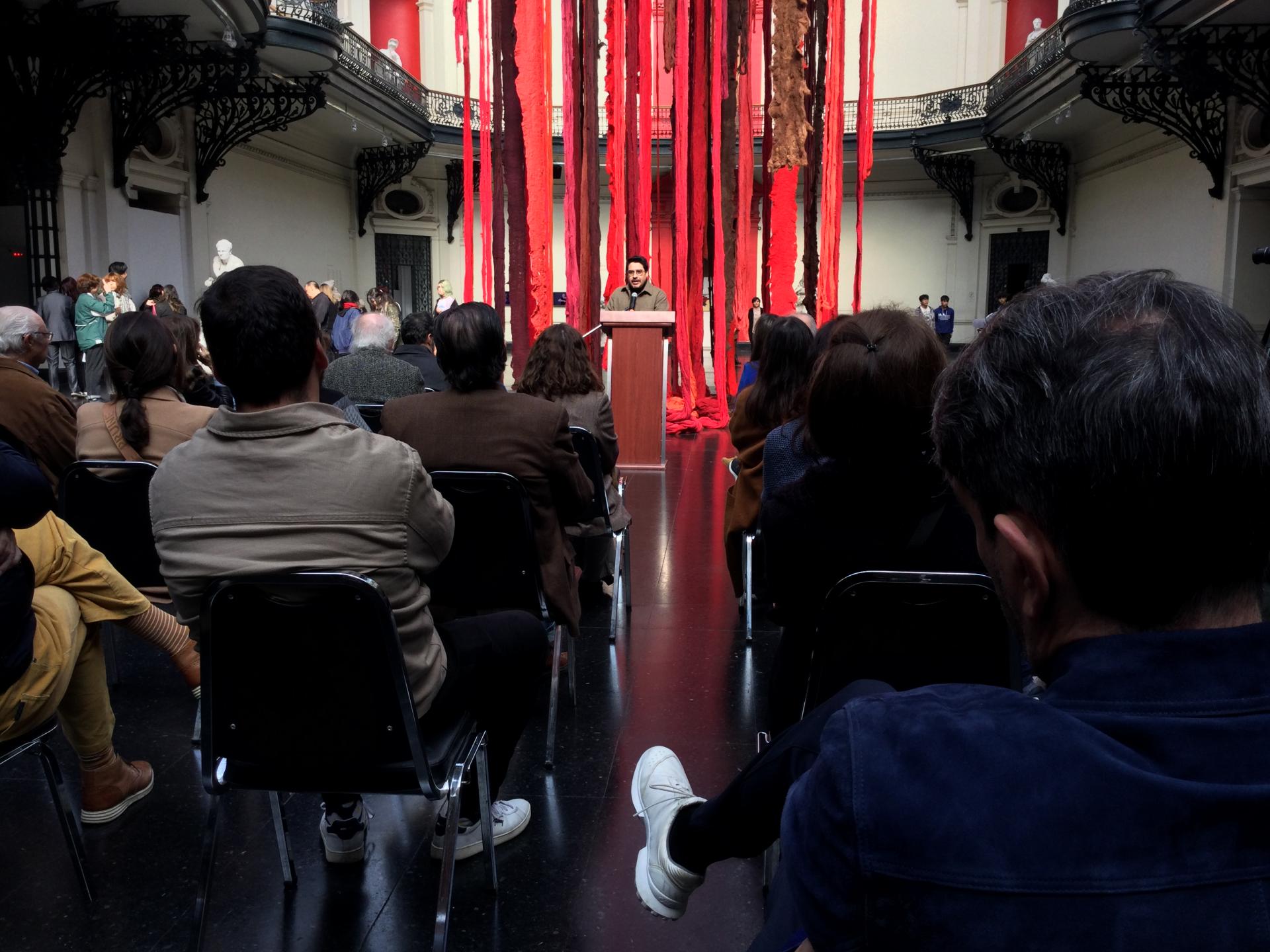 El curador Sebastián Vidal Valenzuela dando un discurso, detrás de él se pueden ver telas colgadas de colores rojo y café. Frente a él hay personas sentadas observandolo.
