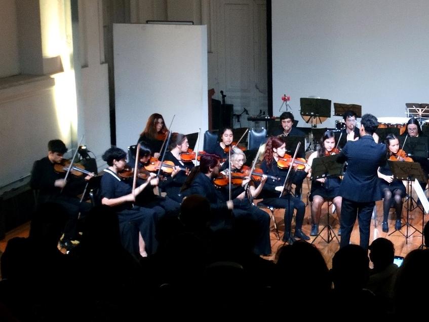 Orquesta tocando sus instrumentos. En primer plano se ven las siluetas del público