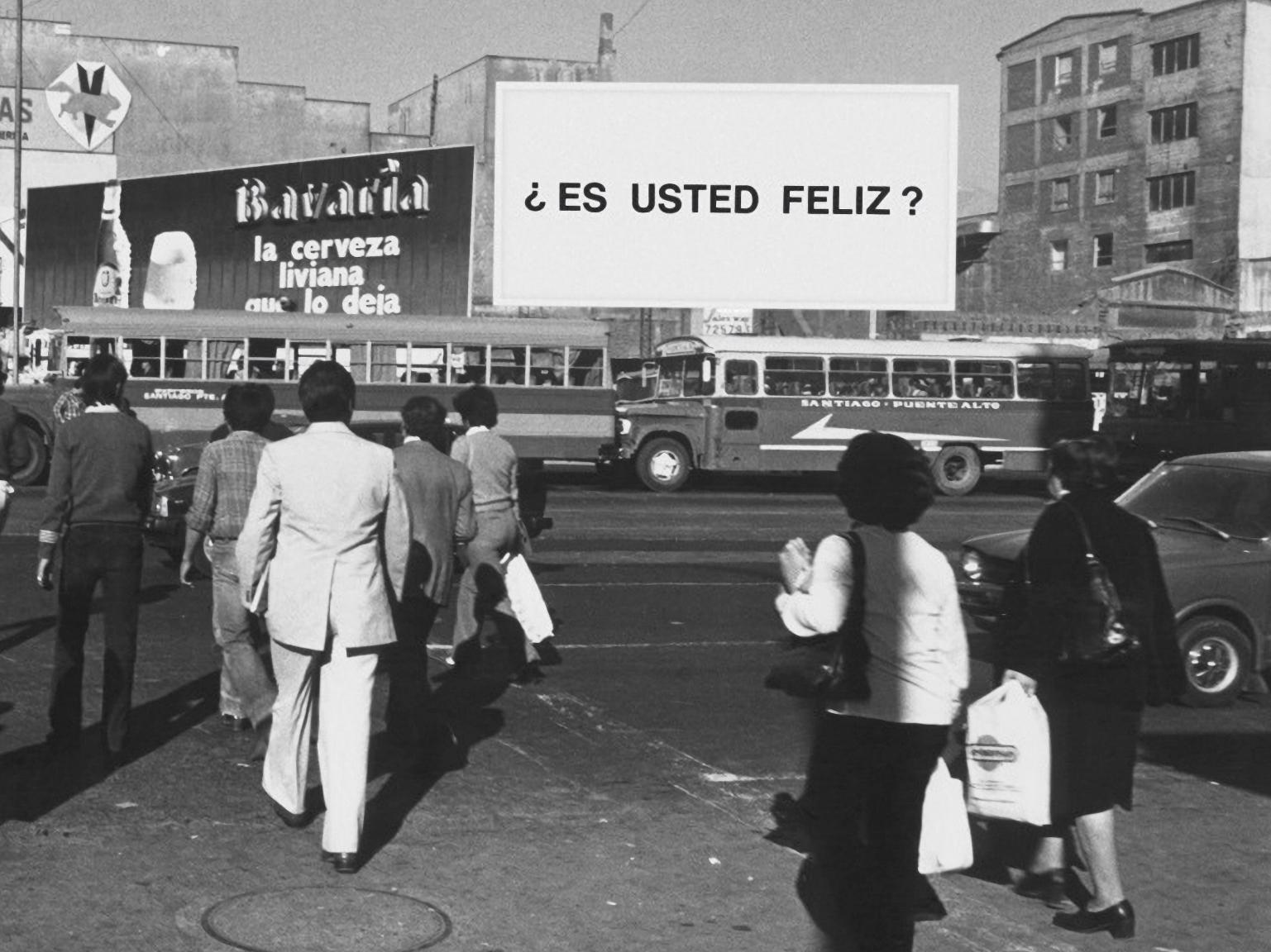 Fotografía en blanco y negro de personas caminando por la calle. Al fondo se visualiza un letrero con la pregunta ¿Es usted feliz?