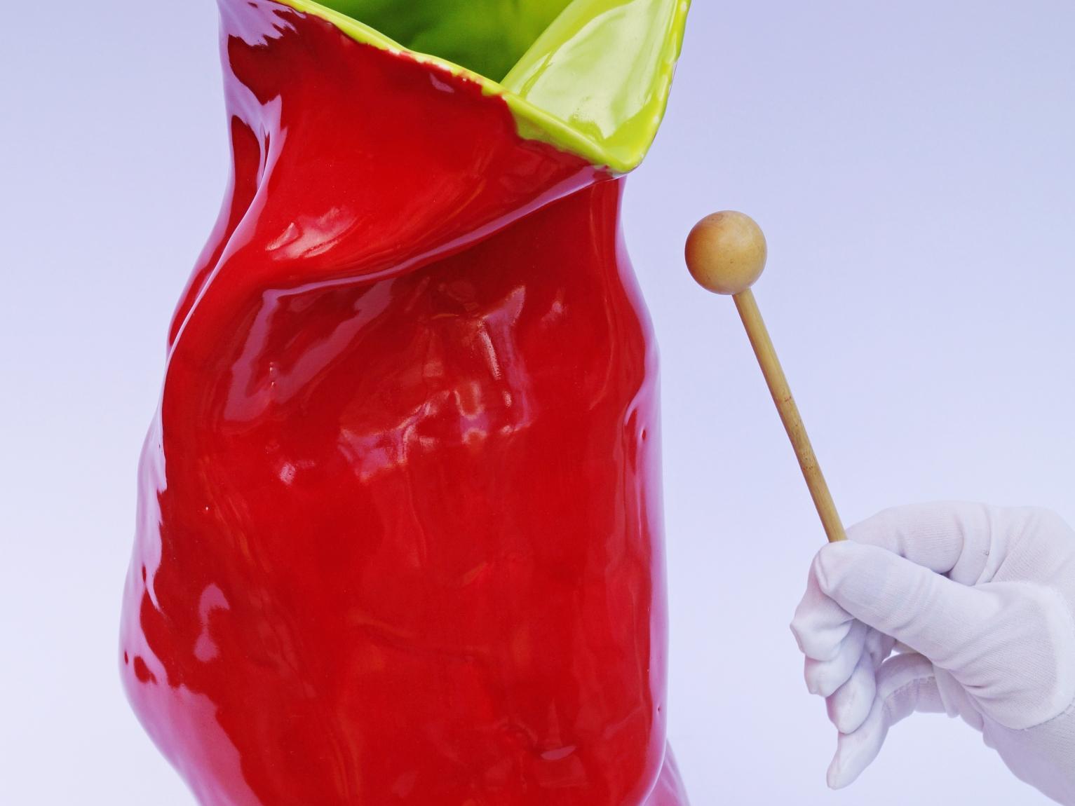 Fotografía de un jarrón cerámico de colores rojo y verde, junto a una mano con un guante blanco y una vara de percusión