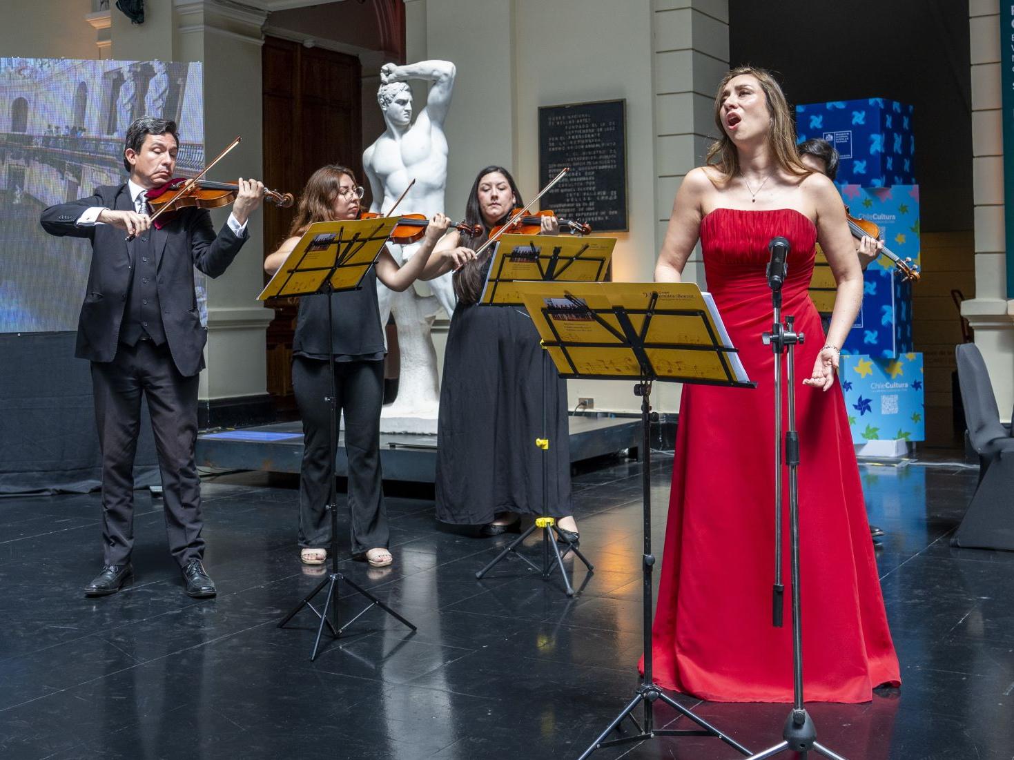 Fotografía de una mujer con vestido rojo, cantando, y tres músicos detrás de ella