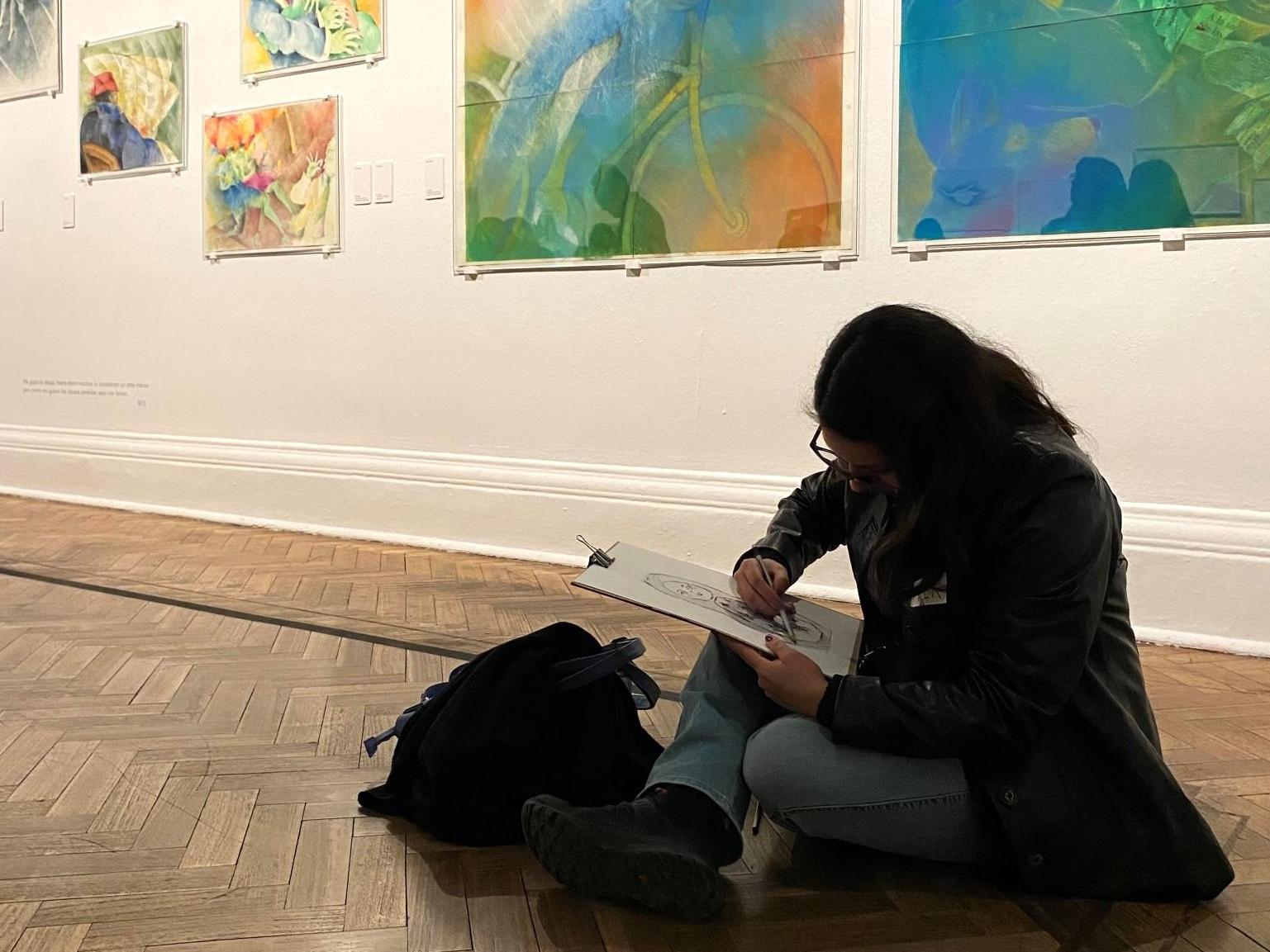 Mujer dibujando, sentada en el piso de una sala de exposición.