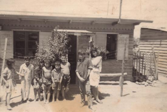 Foto en tonos sepia que muestra a un grupo de niños frente a una casa pequeña de madera