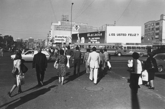 Fotografía en blanco y negro de personas caminando por la calle, con letreros publicitarios de fondo. 
