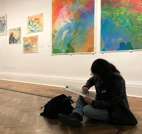 Mujer dibujando, sentada en el piso de una sala de exposición.