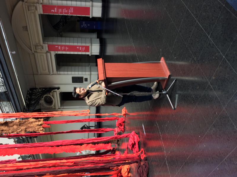 El curador Sebastián Vidal Valenzuela dando un discurso, detrás de él se pueden ver telas colgadas de colores rojo y café. Frente a él hay personas sentadas observandolo.